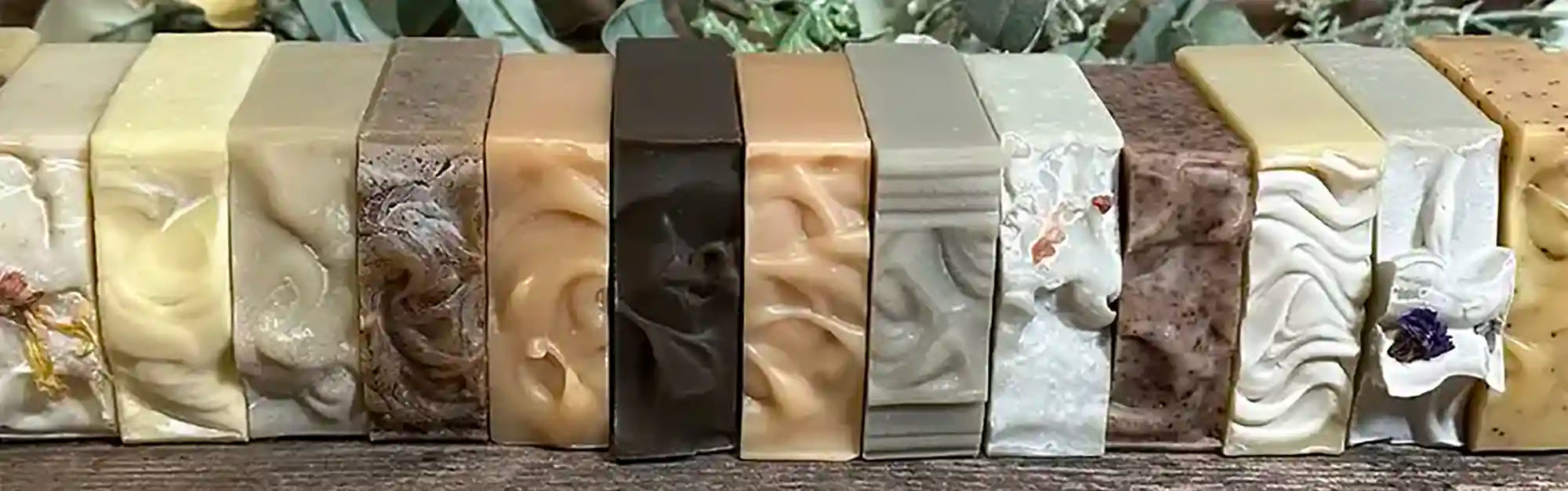 A row of artisanal handmade soaps in various colors and textures, displayed side-by-side on a dark wooden surface with green foliage in the background.