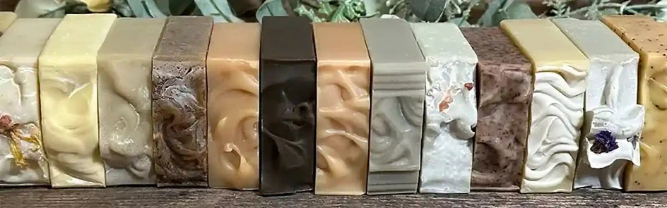 A row of artisanal handmade soaps in various colors and textures, displayed side-by-side on a dark wooden surface with green foliage in the background.